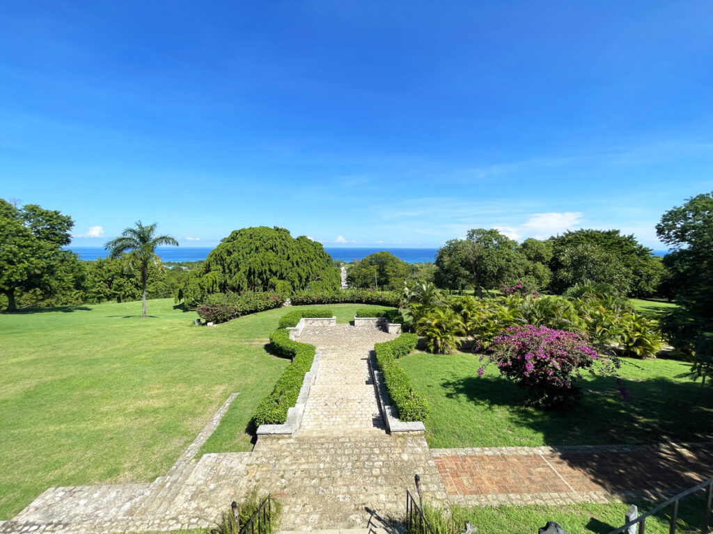 View of the front grounds at Rose Hall Great House in Montego Bay, Jamaica, featuring stone steps, tropical gardens, and a distant ocean backdrop