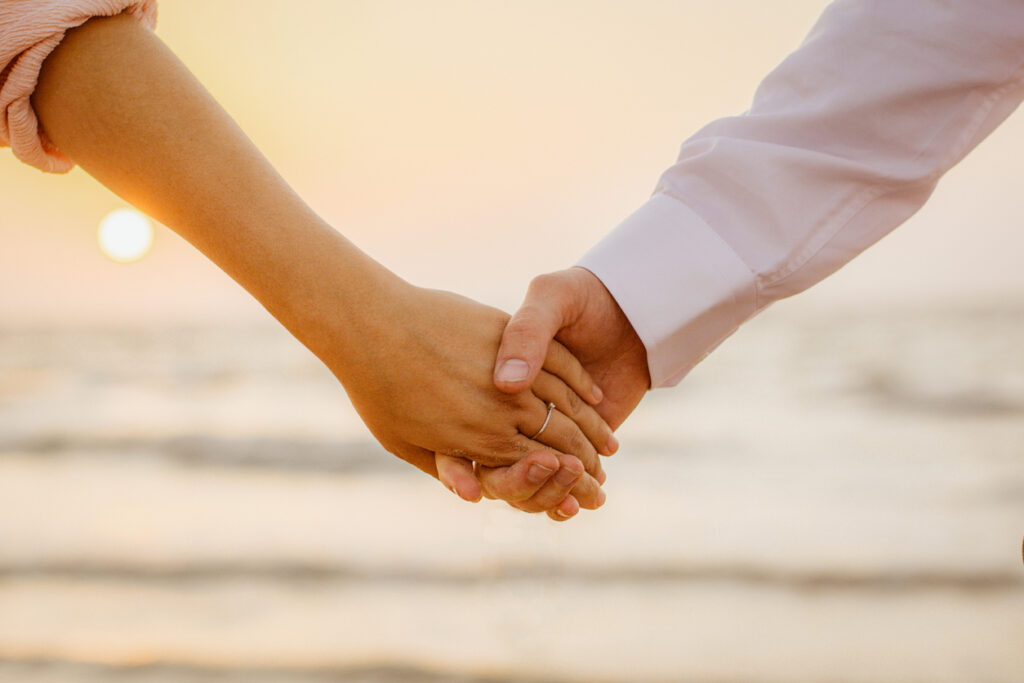 Close‑up of two people holding hands with the ocean in the background, symbolizing love and connection during an anniversary celebration cruise and the meaningful option of renewing vows at sea.