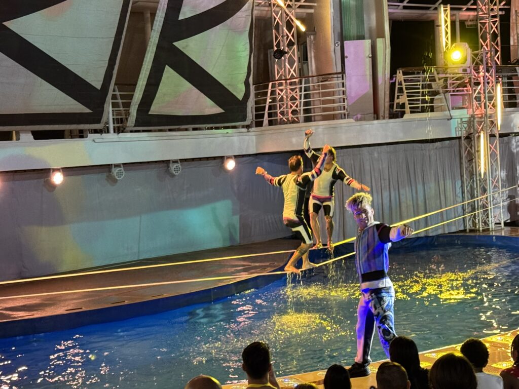 Acrobats performing over the AquaTheater pool during the Oasis of the Seas aqua show, with two performers mid‑air above the slackline and vibrant stage lighting illuminating the scene.