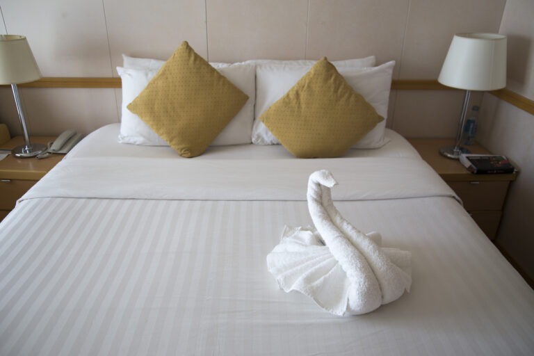 Neatly made cruise cabin bed with white linens, mustard‑yellow pillows, and a towel swan centered on the bed, creating a calm and welcoming stateroom atmosphere.