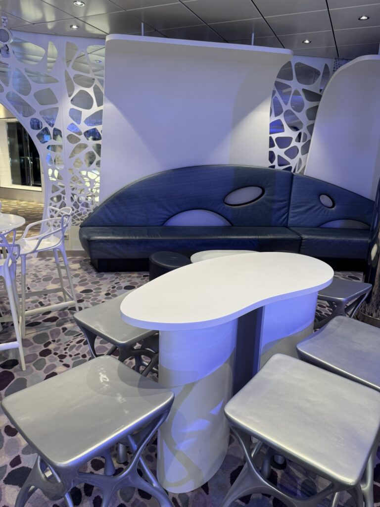 Futuristic seating area at the Bionic Bar on Oasis of the Seas, featuring sculptural silver stools, a curved white table, and a modern blue bench framed by abstract cutout wall panels.