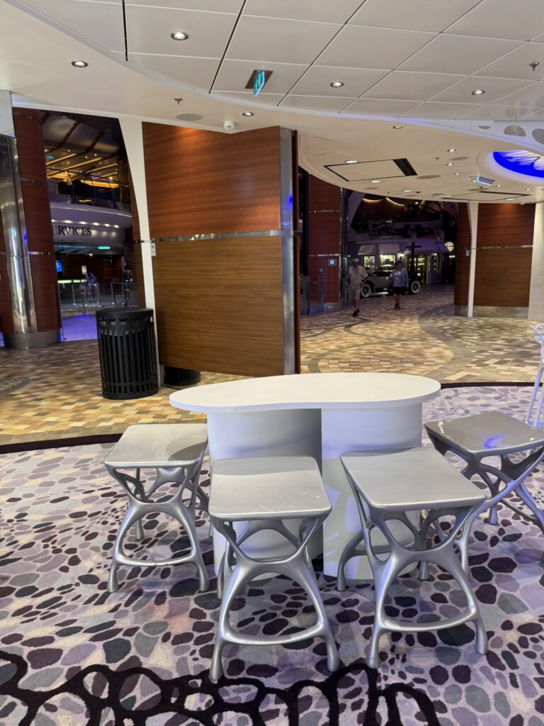 Modern seating area near the Bionic Bar on Oasis of the Seas, featuring sculptural silver stools, a curved white table, patterned flooring, and a view of the ship’s interior walkway with a vintage car display in the distance.