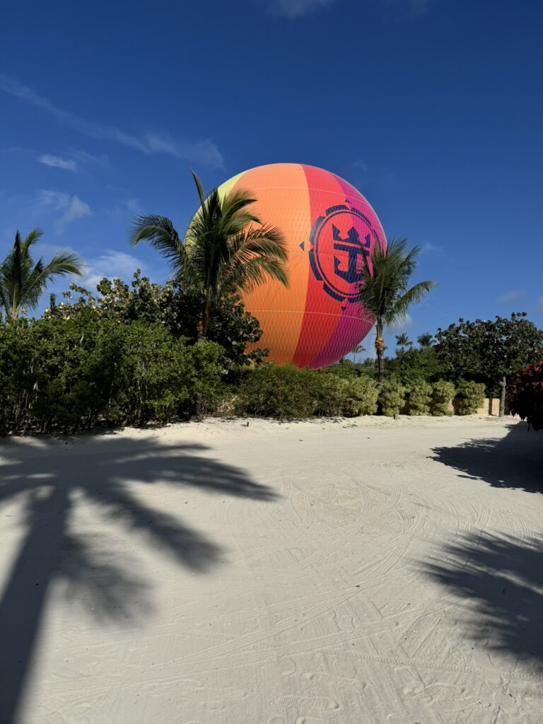 Hot air balloon at Perfect Day at CocoCay featuring the Royal Caribbean logo, rising above tropical palm trees and white sand on a sunny day.