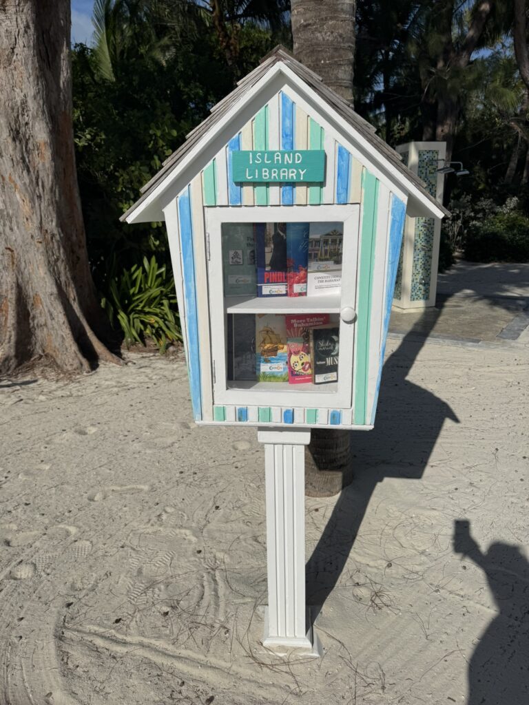 A small, colorful Island Library on CocoCay filled with books, set on sandy ground among tropical trees and greenery.