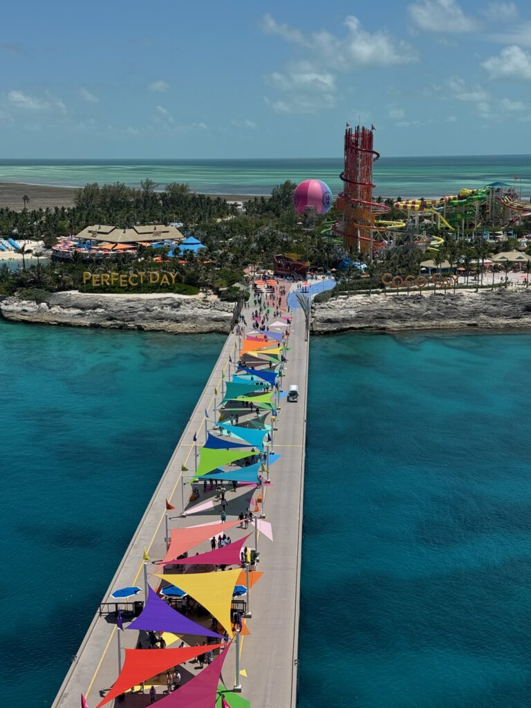 The pier at CocoCay stretching over bright turquoise water, with the island’s palm trees and colorful buildings visible in the distance.