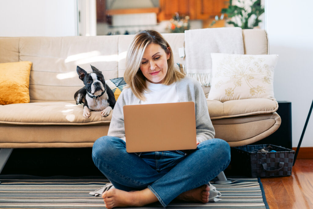 Woman at home using her computer with a Boston Terrier beside her on the couch, reviewing cruise details and completing important planning steps tied to cruise mistakes to avoid, such as making reservations, checking in online, and organizing pre‑cruise tasks.