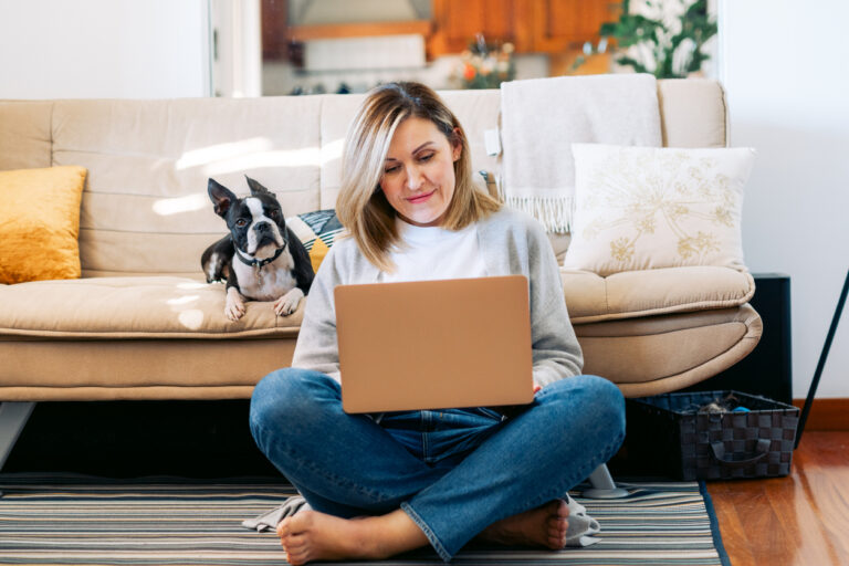 Woman at home using her computer with a Boston Terrier beside her on the couch, reviewing cruise details and completing important planning steps tied to cruise mistakes to avoid, such as making reservations, checking in online, and organizing pre‑cruise tasks.