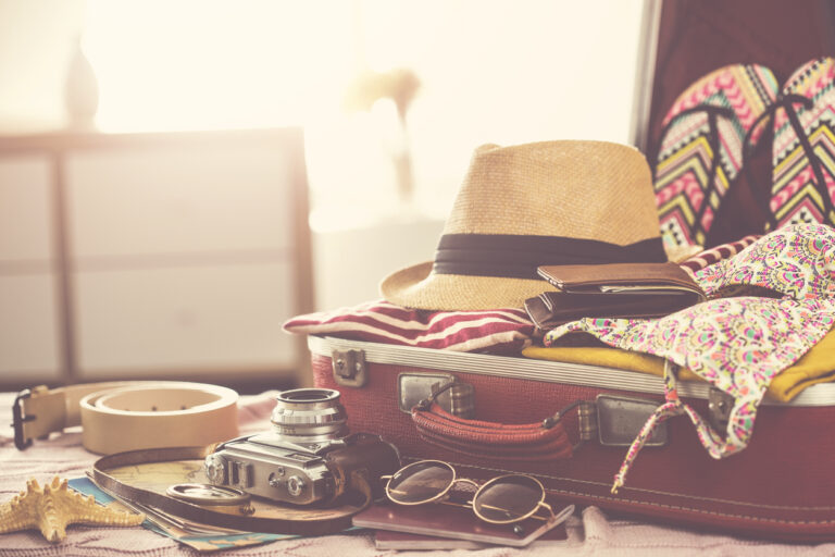Open suitcase surrounded by cruise vacation essentials, including clothing, flip‑flops, a passport, sunglasses, and a straw hat, arranged for a cruise packing list.