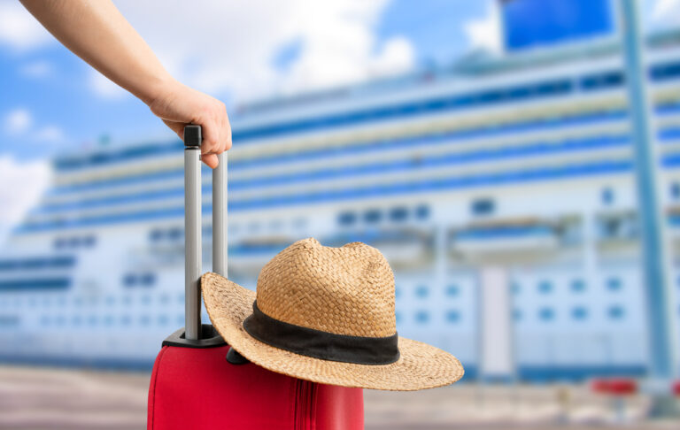 Close‑up of a red suitcase with a straw hat on top, pulled by a traveler with a large cruise ship blurred in the background.