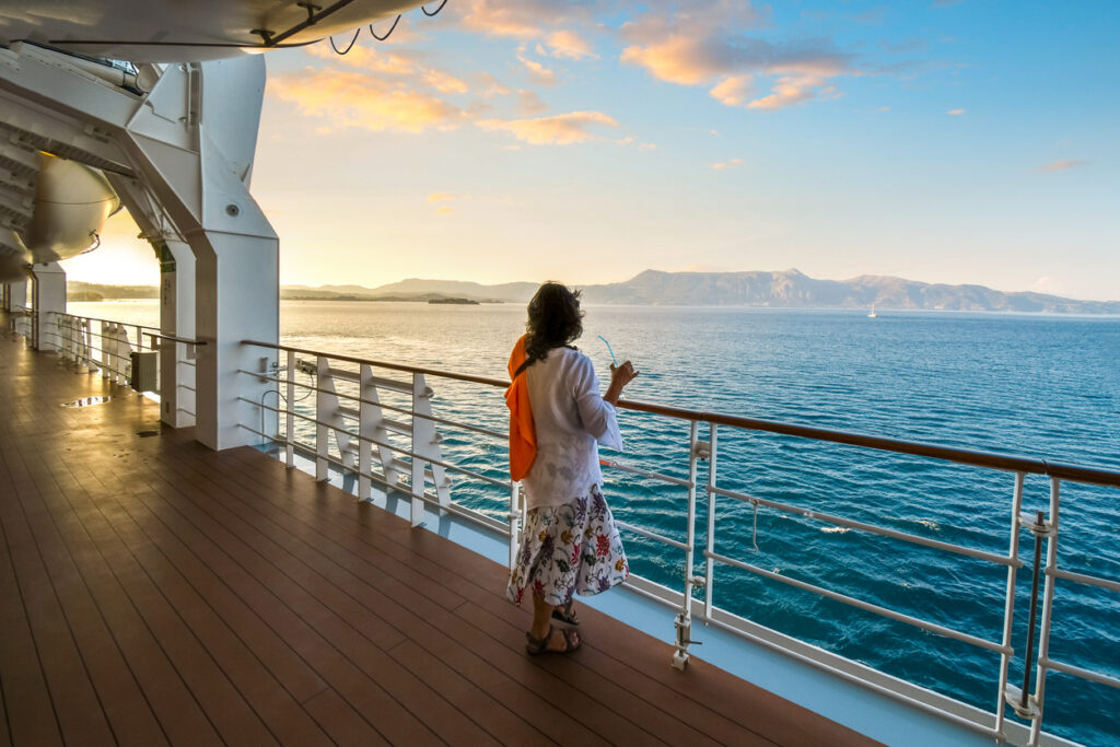 Person standing on a cruise ship deck at sunset, looking out at the ocean and distant mountains, capturing the peaceful experience highlighted in first time cruiser tips.