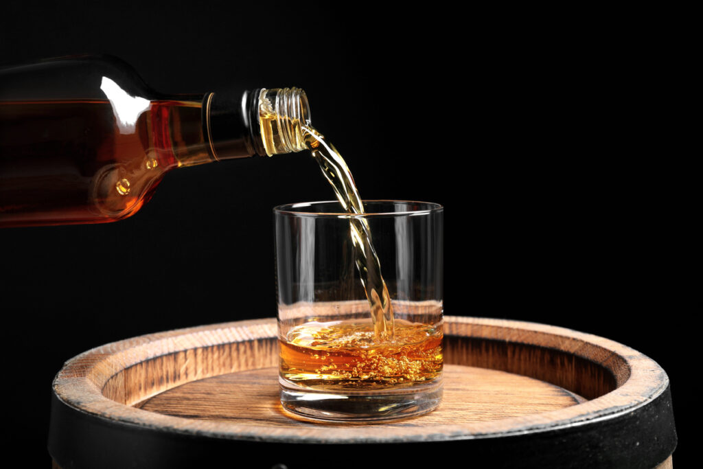 Golden rum being poured from a bottle into a glass resting on a wooden barrel, creating a warm and rustic distillery feel