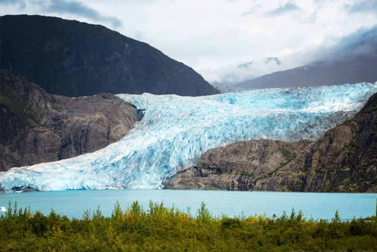 A massive blue‑and‑white glacier flowing down a rocky mountainside into a turquoise lake, surrounded by green vegetation and misty clouds.