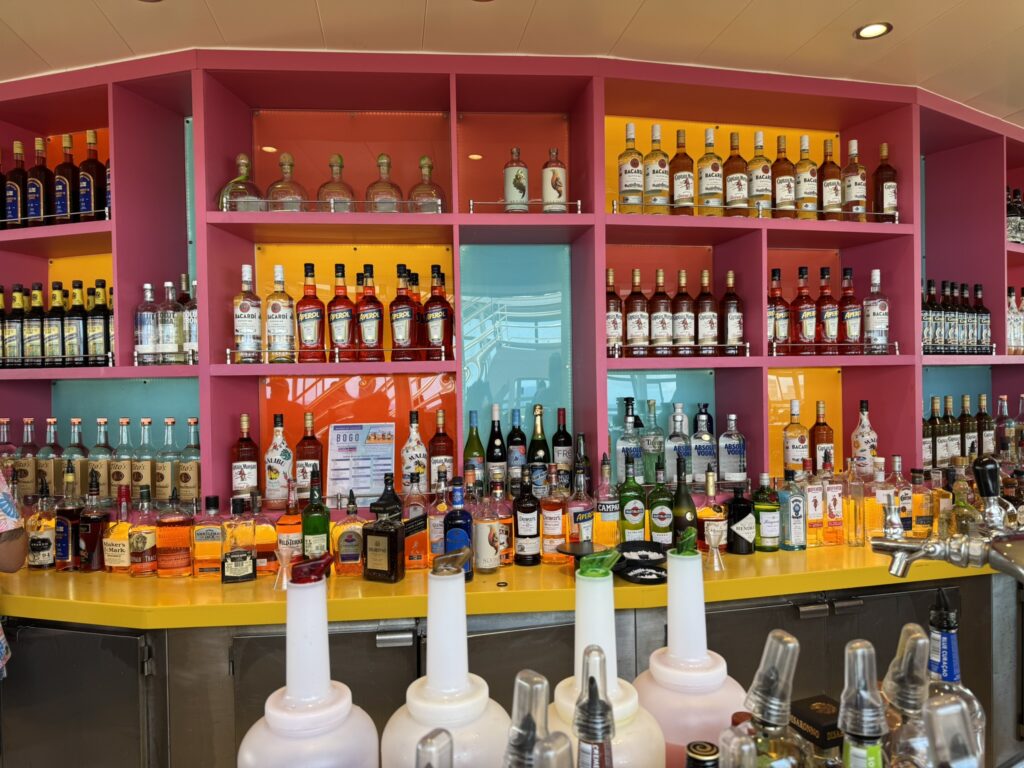 Poolside Lime & Coconut bar on Oasis of the Seas with bright pink and orange shelving filled with liquor bottles and a bartender preparing drinks.