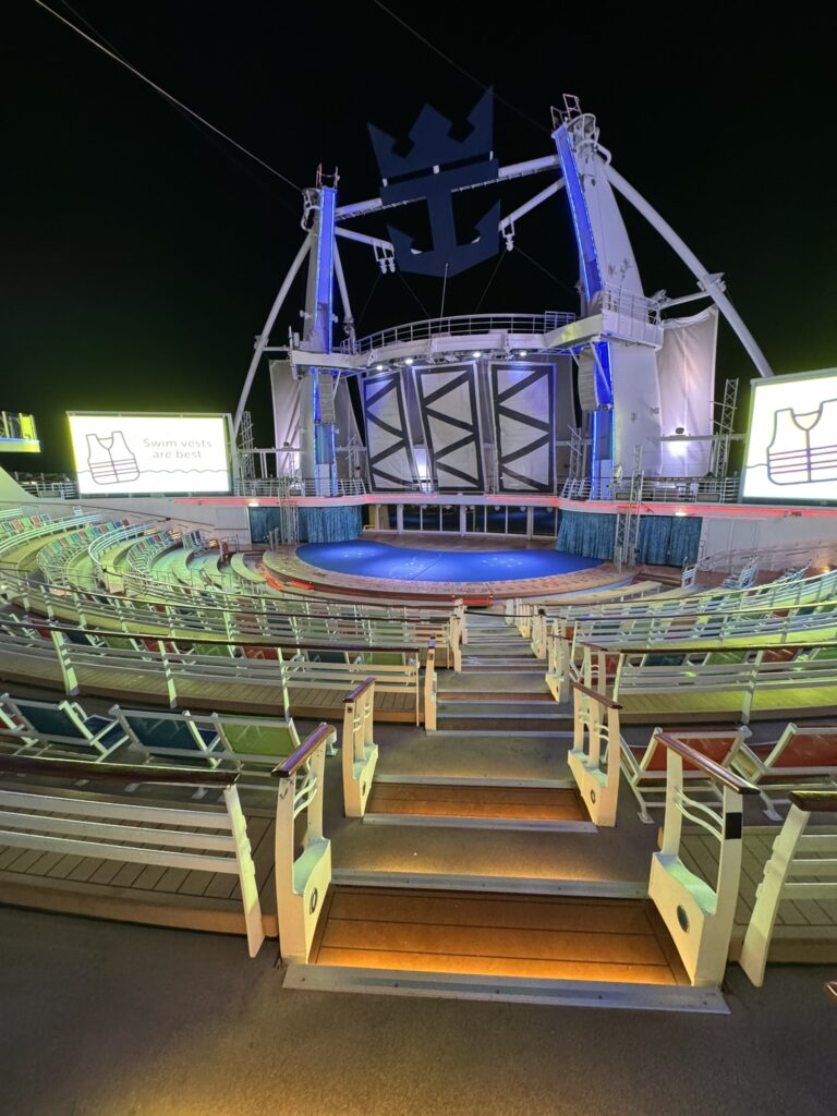 AquaTheater seating area on Oasis of the Seas at night, with curved rows of seats, large digital screens, and the main stage where scheduled movies and game broadcasts take place.