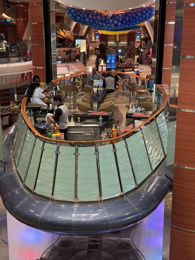 Rising Tide Bar on Oasis of the Seas, showing the floating platform surrounded by tall columns, seating areas, and the open atrium as the bar moves between decks.