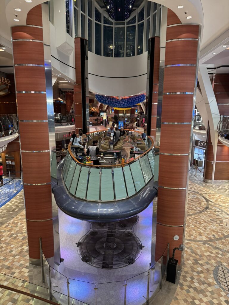Rising Tide Bar on Oasis of the Seas, showing the floating platform surrounded by tall columns, seating areas, and the open atrium as the bar moves between decks.
