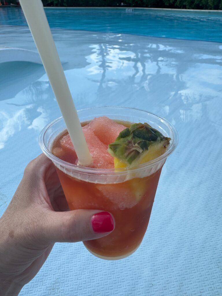 Hand holding a frozen tropical drink garnished with pineapple and watermelon beside a cruise ship pool, illustrating a refreshing option included with the Royal Caribbean drink package.