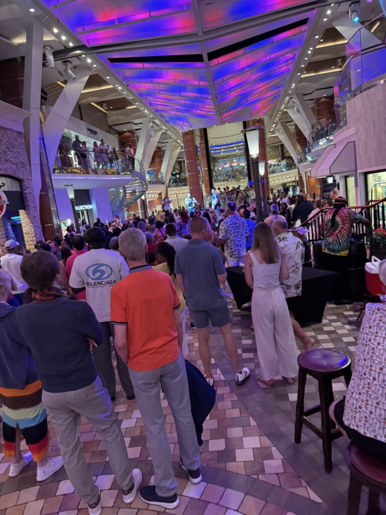 Crowded Royal Promenade on Oasis of the Seas with guests filling the walkway under bright purple lighting during a busy evening event.