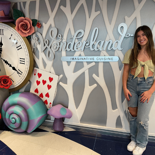 Daughter standing in front of the Wonderland Restaurant on Royal Caribbean’s Wonder of the Seas, surrounded by the whimsical, colorful décor that reflects the imaginative theme of Wonderland Restaurant on Royal Caribbean.