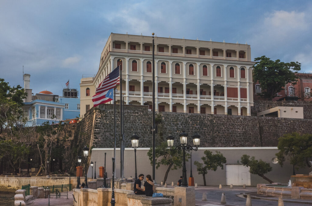 People relaxing along Paseo de la Princesa in Old San Juan, Puerto Rico, with the U.S. and Puerto Rico flags displayed along the scenic waterfront promenade.