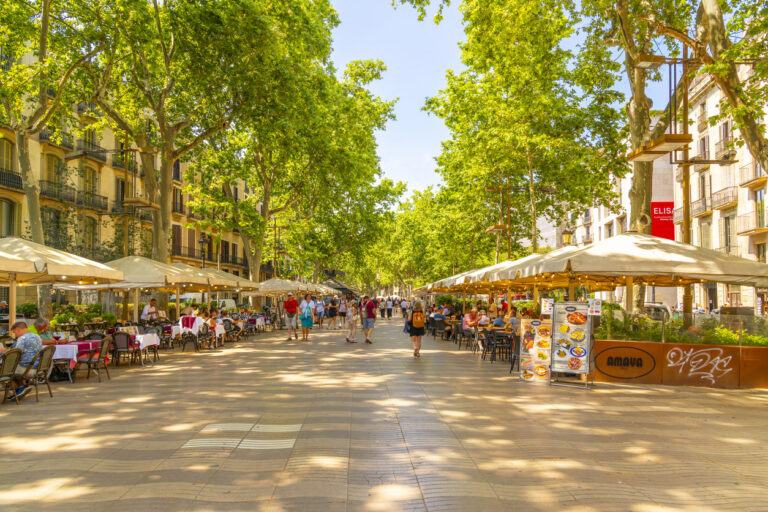 Early morning on the pedestrian Las Ramblas boulevard through historic Barcelona Spain as tourists and locals pass by shops and sidewalk cafes near in the historic Gothic Quarter.