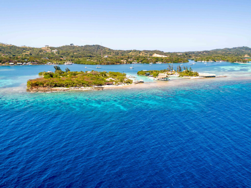 Aerial view of Big French Key and Little French Key off the coast of Roatán, Honduras, surrounded by turquoise Caribbean water, coral reefs, and anchored boats.