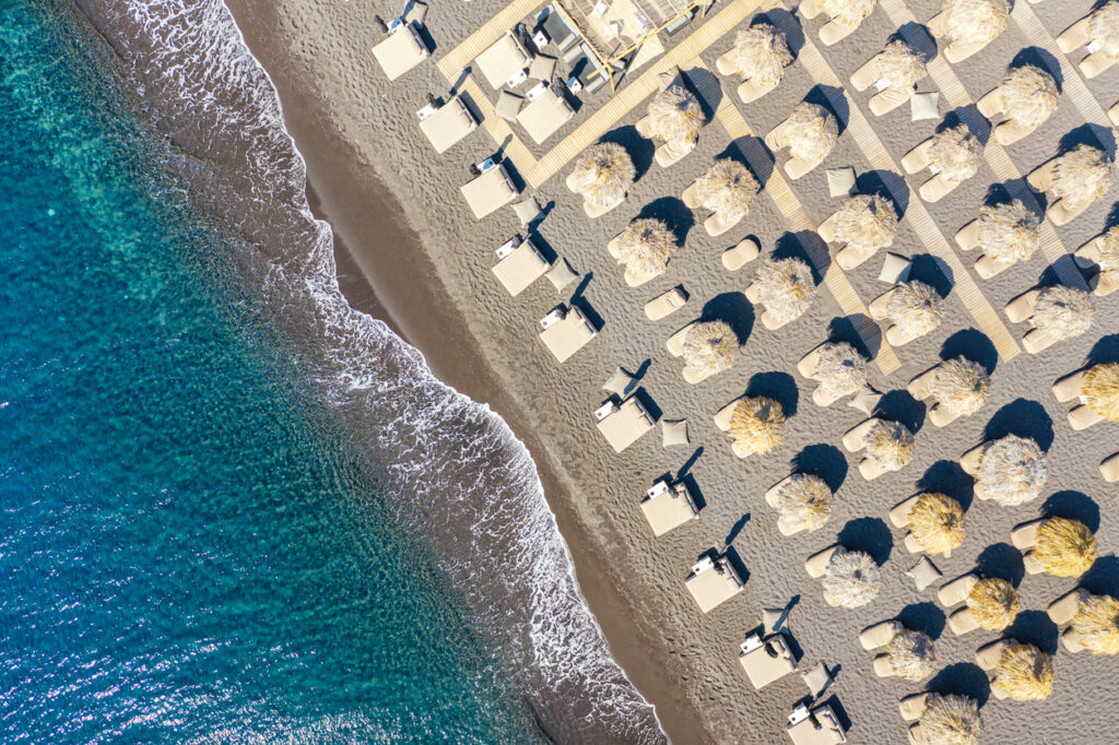 Top view aerial drone photo of black Perissa beach with beautiful turquoise water, sea waves and straw umbrellas. Vacation travel background. Aegean sea, Santorini Island, Greece.