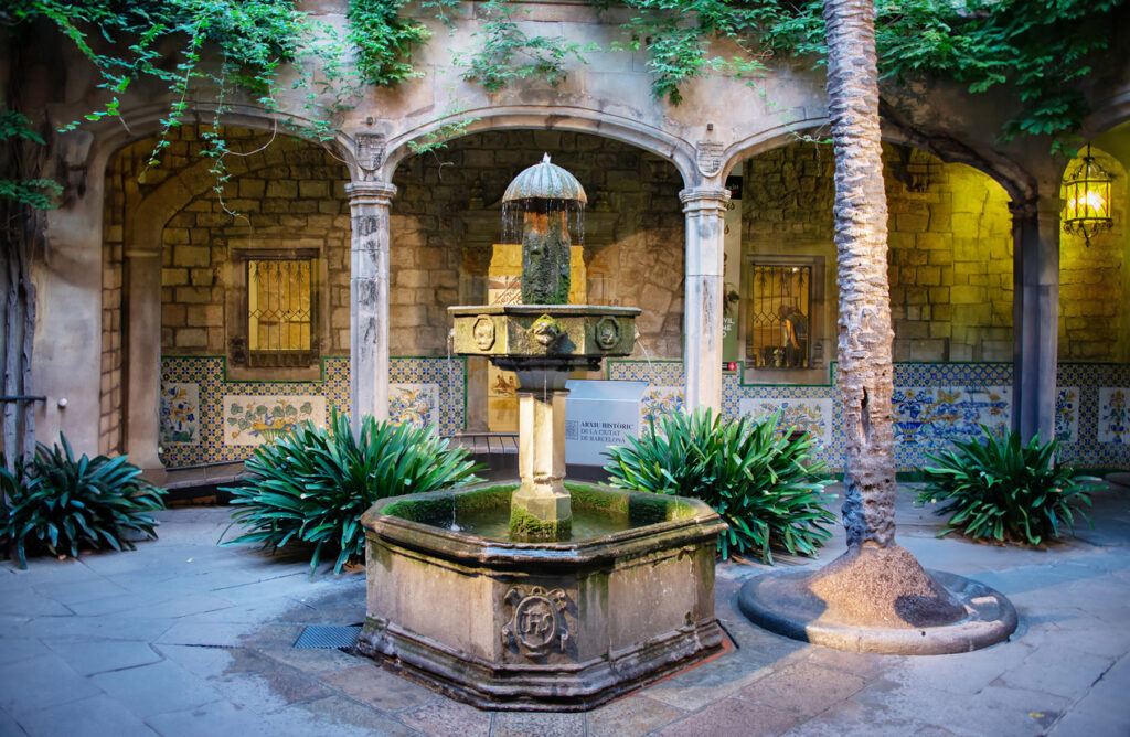 Barcelona, Spain - A photo of an old, octagonal fountain surrounded by arched columns, green foliage, decorative tiles and interesting wall lamps in the Picasso Museum on a summer morning in the Gothic Quarter of Barcelona, Spain.