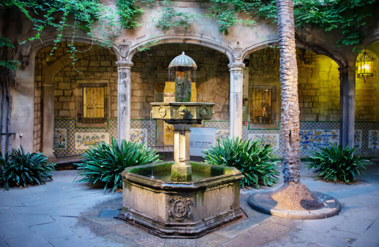 Barcelona, Spain - A photo of an old, octagonal fountain surrounded by arched columns, green foliage, decorative tiles and interesting wall lamps in the Picasso Museum on a summer morning in the Gothic Quarter of Barcelona, Spain.