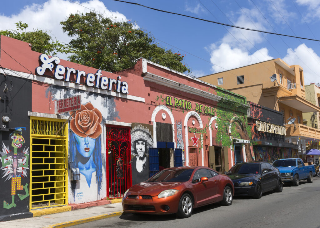 Street scene in the Tras Talleres area of Santurce, San Juan, Puerto Rico, featuring colorful wall art, bars, restaurants, and parked cars under a bright blue sky.