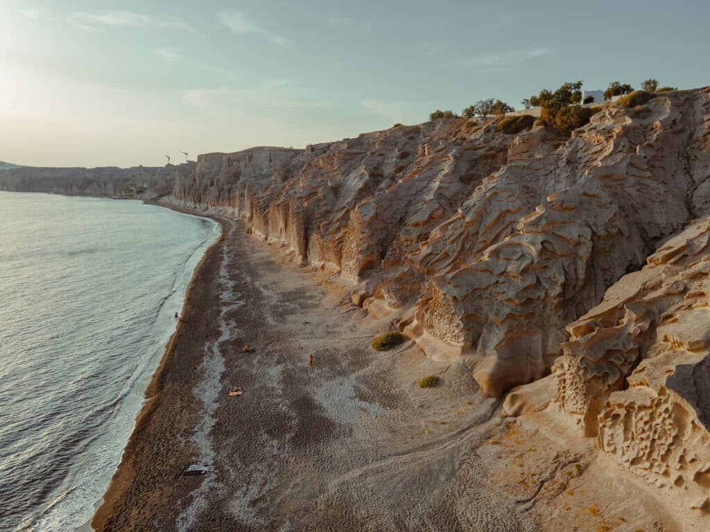 Aerial view gliding alongside the rocky cliffs and shimmering blue waters of the Mediterranean Sea at a Santorini beach, Greece.