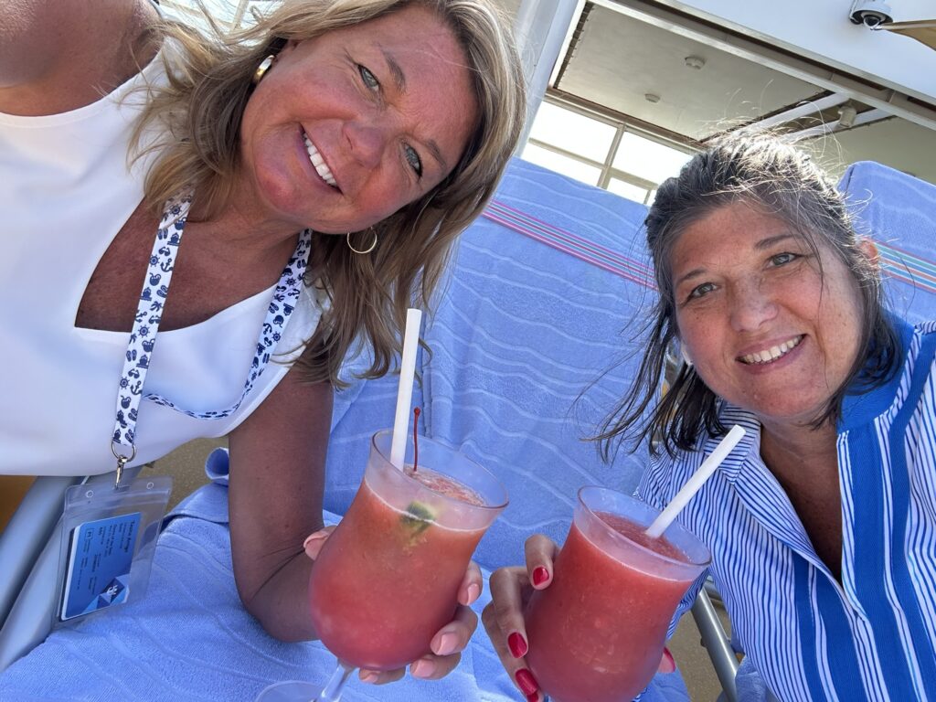Two people relaxing on pool deck lounge chairs with drinks, enjoying their cruise ship drink package without worrying about the bar tab.