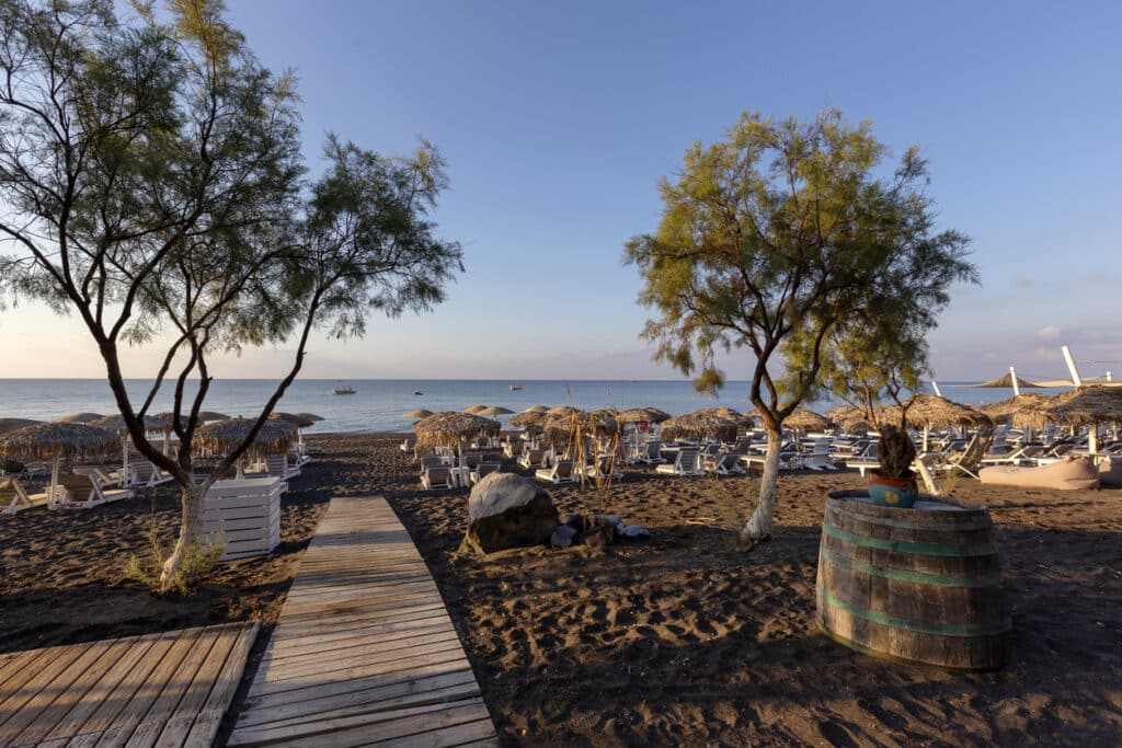 Santorini black beach Perivolos illuminated by the morning rays of the sun. Beach chairs with umbrellas. Two olive trees. Beautiful calm sea.