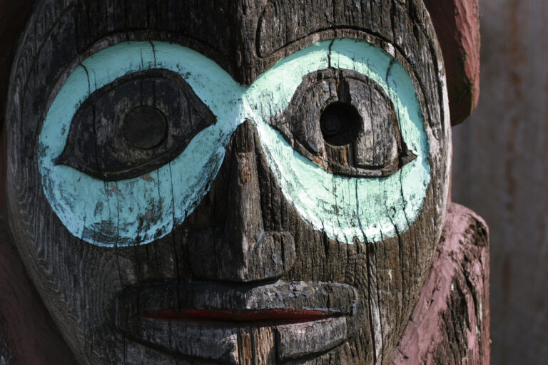 Close‑up of a carved wooden totem figure displayed in a museum setting, showing detailed facial features and painted accents.