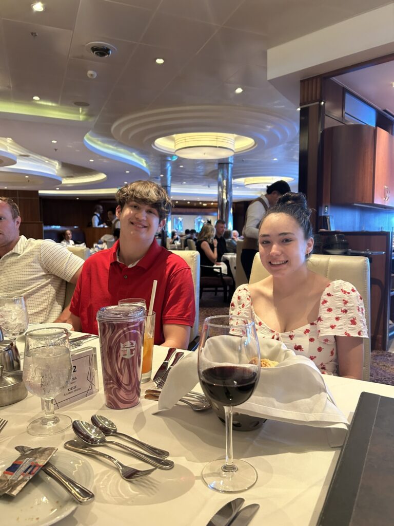 Guests enjoying a seated dinner in the main dining room, with a set table, warm lighting, and a relaxed cruise atmosphere.