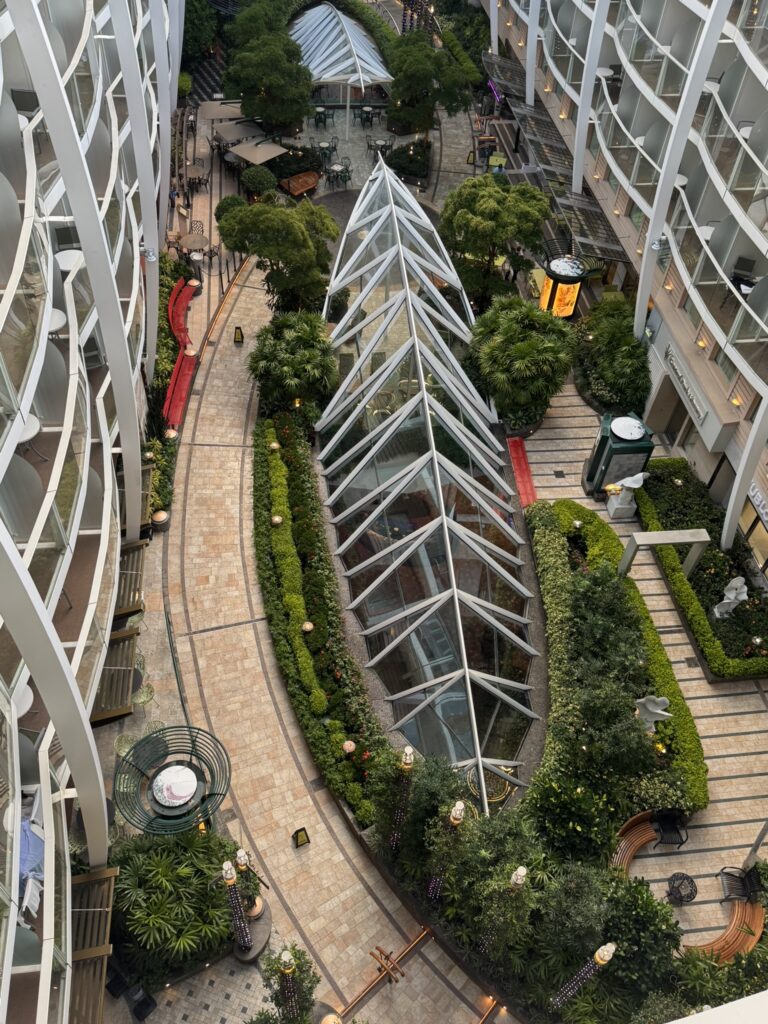 Open‑air central courtyard on a cruise ship with lush greenery, curved walkways, and balconies overlooking the space.