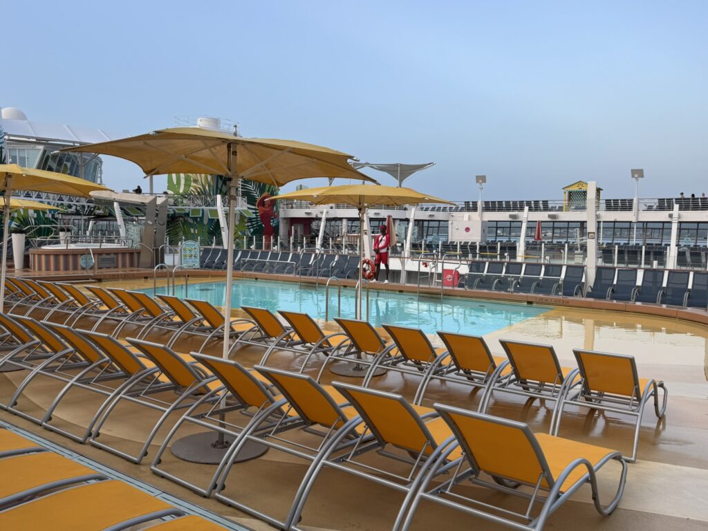 Cruise ship pool deck with rows of yellow loungers, umbrellas, and a central swimming pool under clear skies.