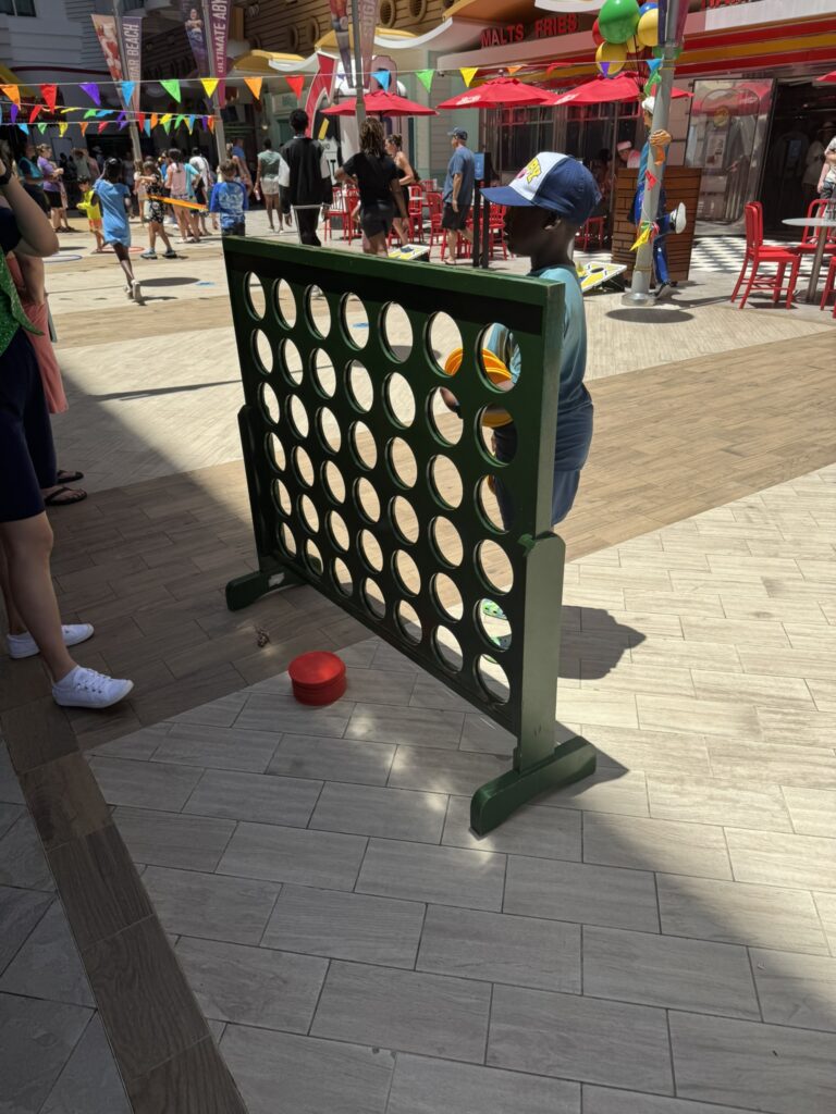 A child plays a giant Connect Four game in a lively outdoor area decorated with colorful pennant banners, showing one of the many free activities available onboard.