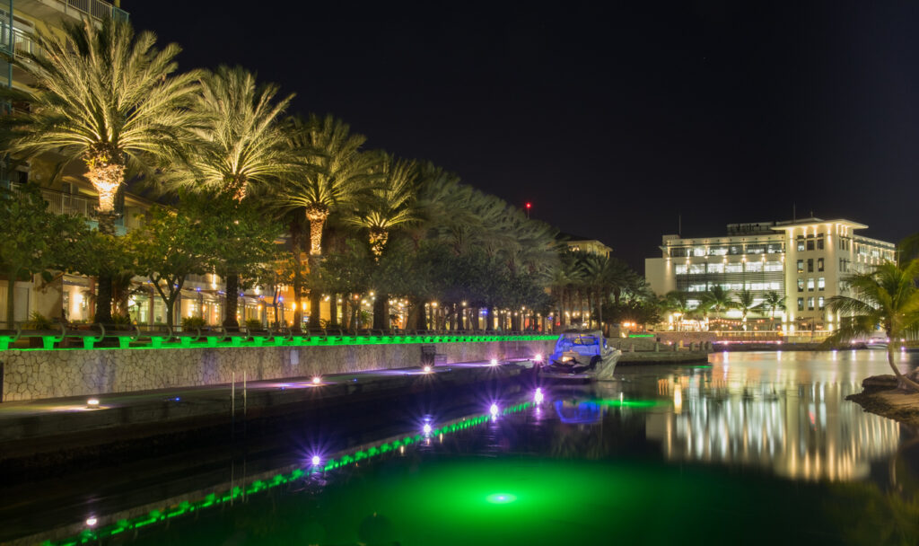 Nighttime view of the Camana Bay marina in Grand Cayman with a yacht moored to a colorful, illuminated pier