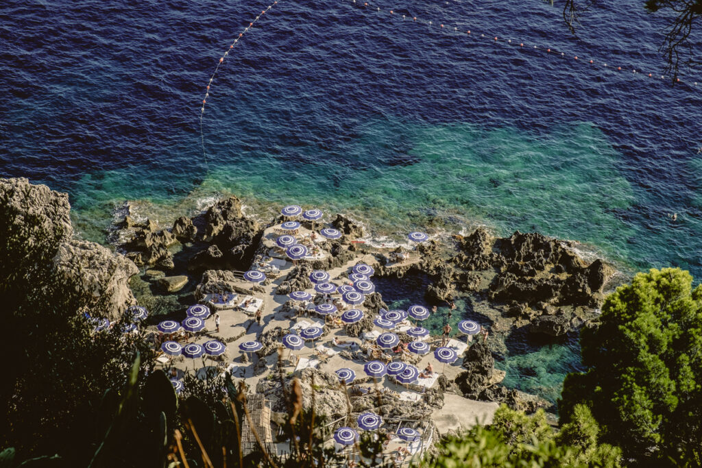 Aerial view of La Fontelina Beach Club on Capri with blue umbrellas, rocky terraces, and turquoise Mediterranean water along the Amalfi Coast