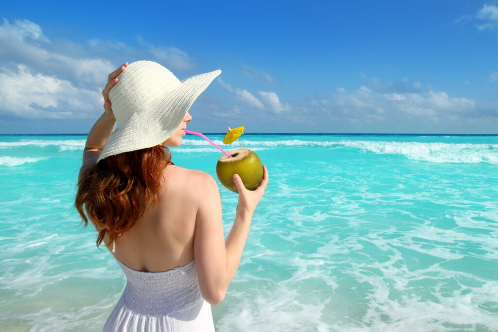 Woman in a beach hat enjoying a fresh coconut drink while standing in calm Caribbean water, capturing the relaxed vibe often found at Costa Maya beach clubs.