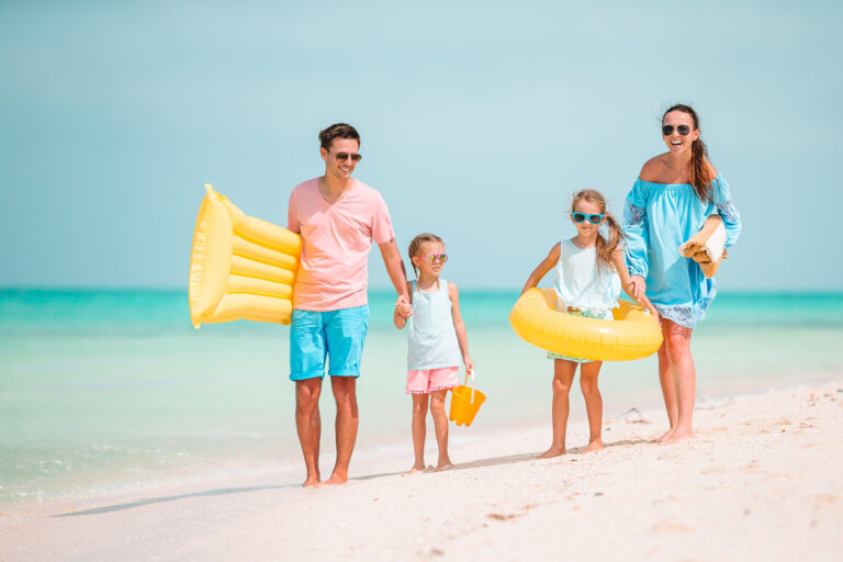 Family walking along a sandy tropical beach with inflatables and beach gear on a sunny day.