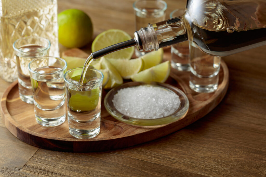 Shot glasses of tequila being poured on a wooden tray with lime wedges and salt.