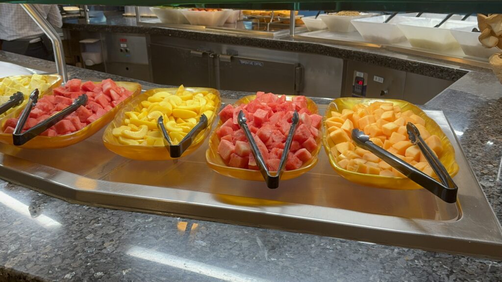resh fruit options at a cruise ship buffet, including pineapple, watermelon, peaches, and cantaloupe in self‑serve trays.