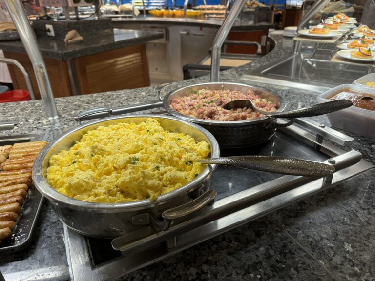 Breakfast options displayed at a cruise ship buffet, including scrambled eggs, corned beef hash, sausages, and small plated dishes.