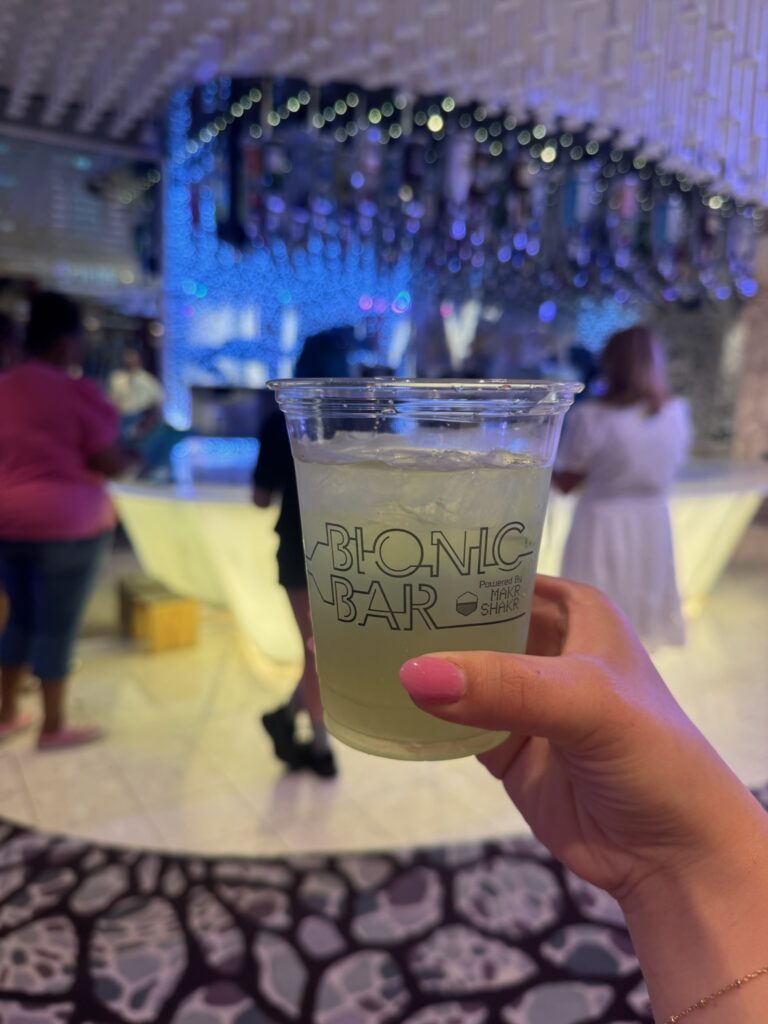Hand holding a cocktail with the Bionic Bar in the background, highlighting the ease of using a cruise ship drink package.