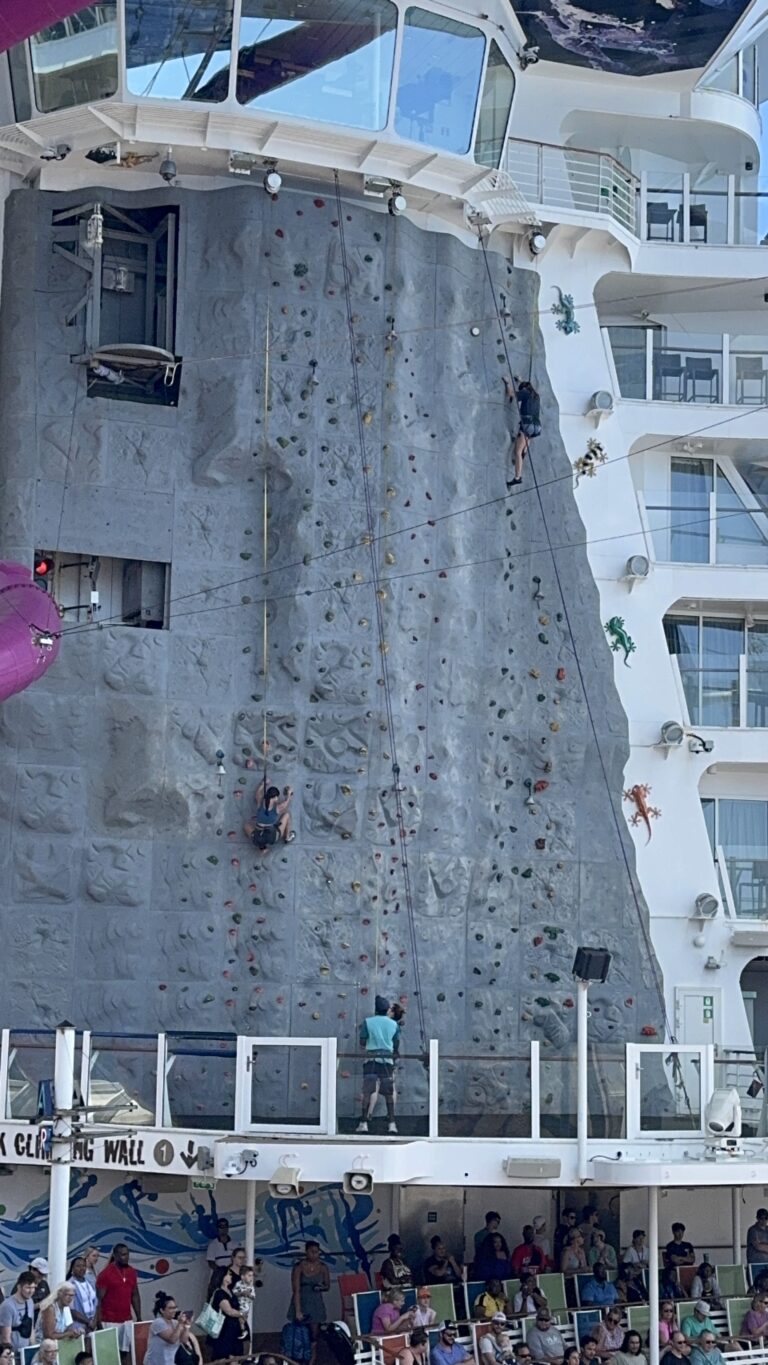 Guests climbing the cruise ship rock‑climbing wall on the upper deck, with multiple climbers on the wall and a crowd watching below.