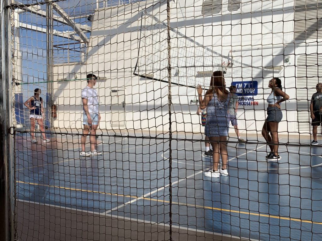 Guests playing volleyball on the cruise ship sports court, with a netted enclosure and bright court markings.