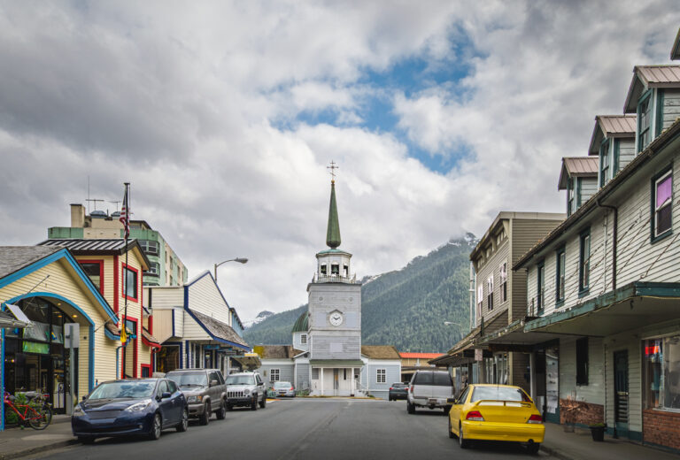 Historic Lincoln Street in downtown Sitka, Alaska, lined with shops and cafés in spring, with St. Michael’s Russian Orthodox Cathedral at the end of the street and mountains rising in the background.