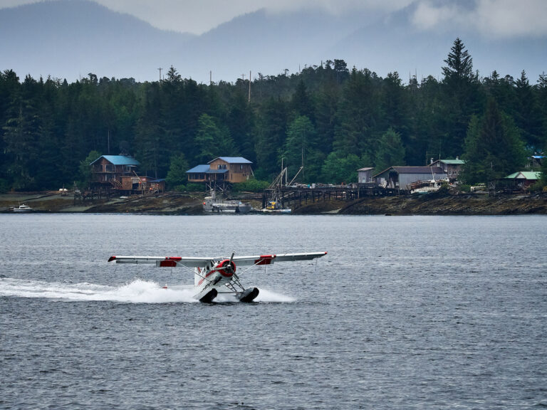 Seaplane landing on calm water near forested shoreline and coastal homes in Ketchikan, Alaska.